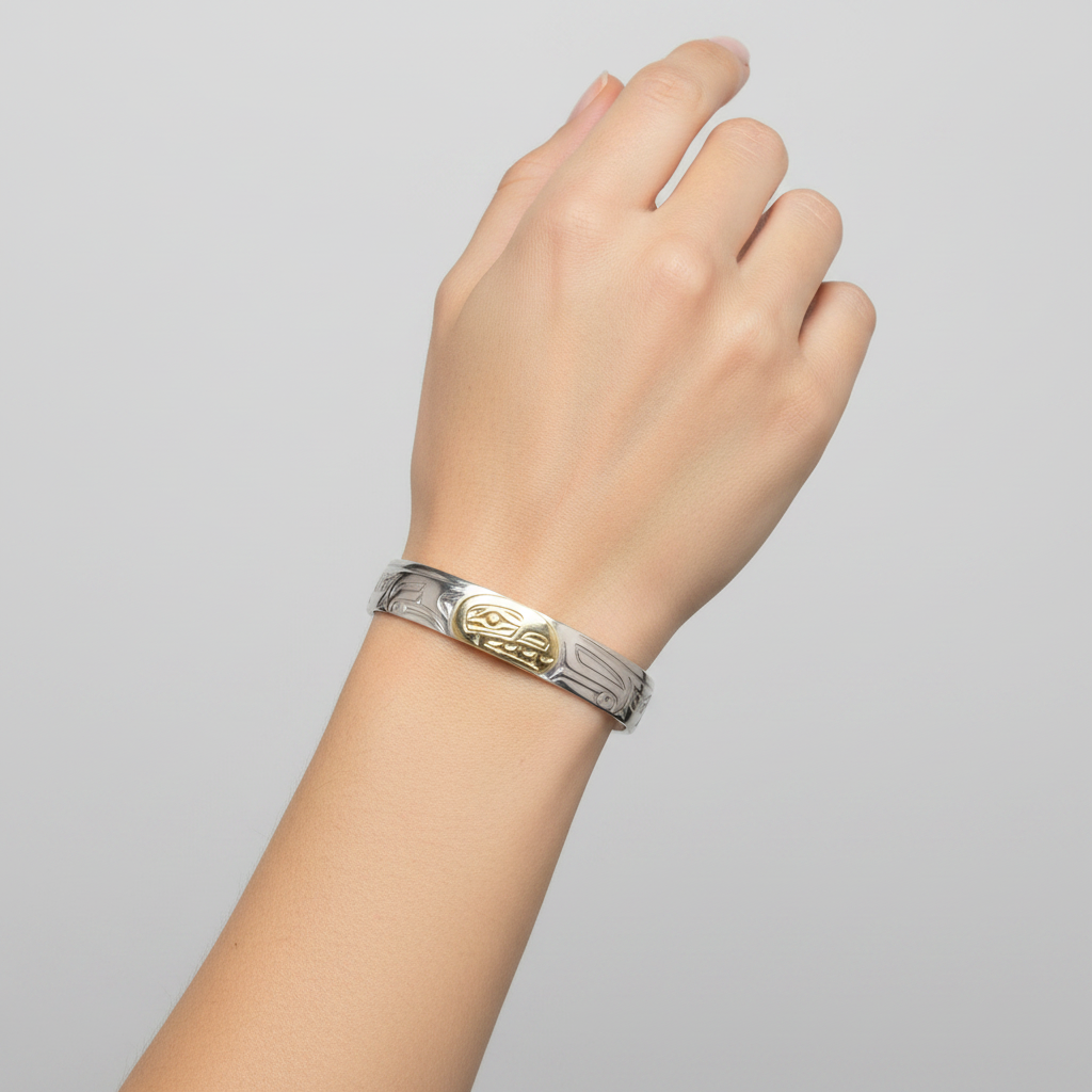 A silver cuff bracelet featuring a polished gold oval centerpiece with a carved indigenous-style face motif, set against a plain white background.