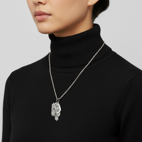 A silver-toned metal pendant featuring a stylized Indigenous-inspired face design with engraved details and a hanging loop at the top, shown on a plain white background.