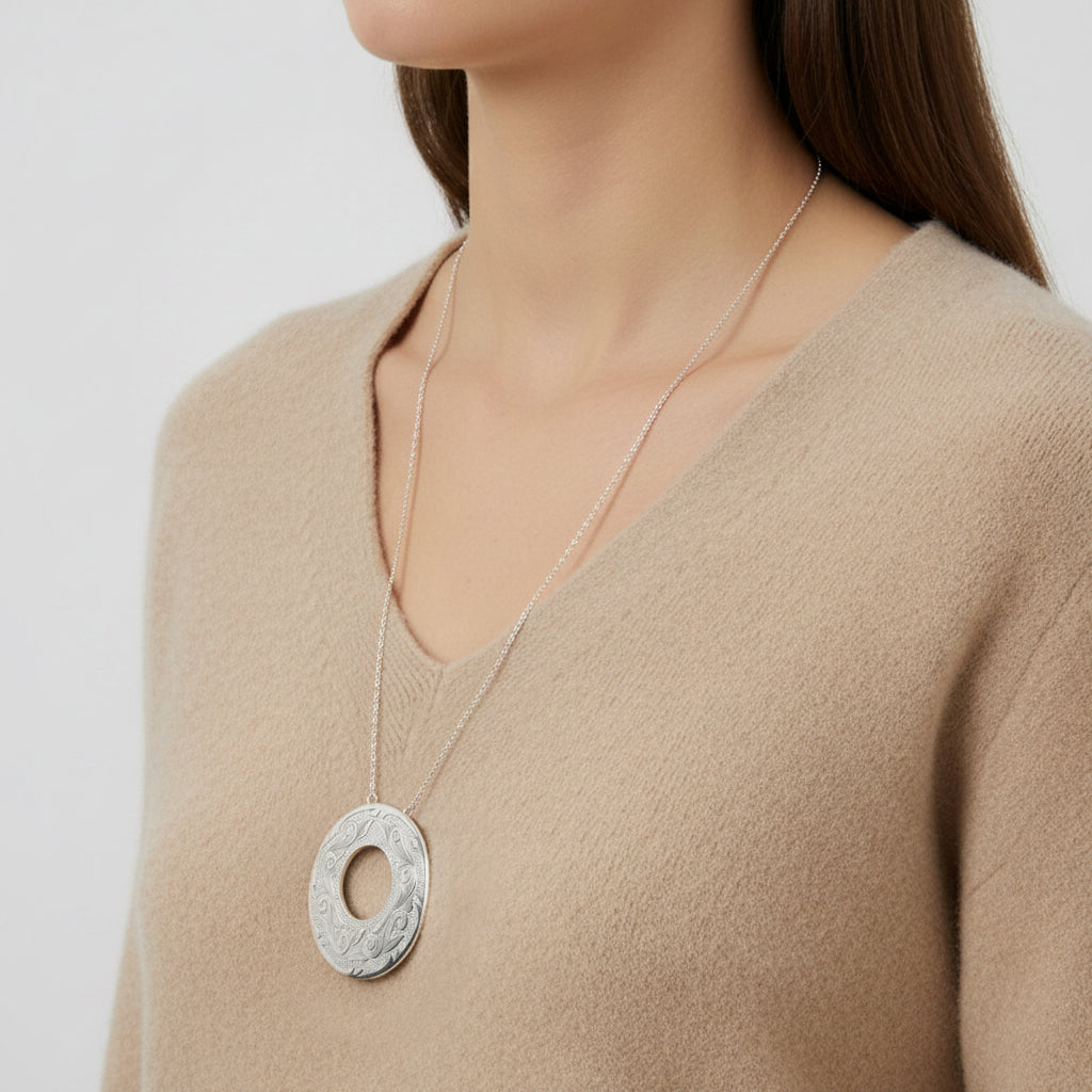 A close-up of a woman wearing a silver pendant necklace with a circular, textured disc design on a delicate chain, paired with a beige knit sweater.